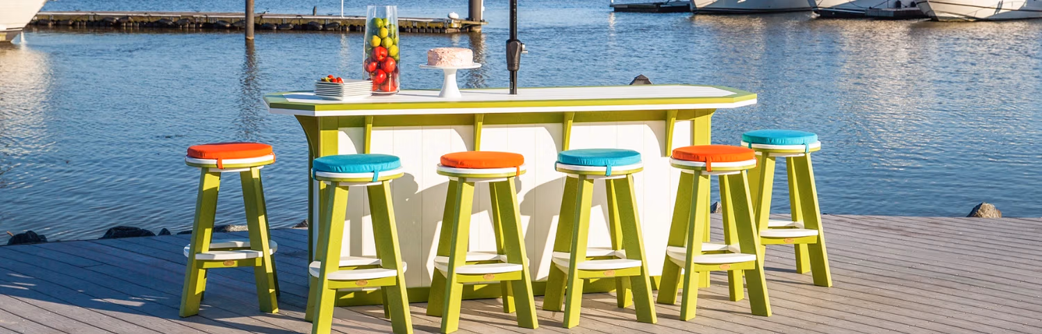 bar-counter-stool-banner.webp A waterside bar on a wooden deck with a white and green counter, six green bar stools with orange and teal cushions, a bowl of apples, a stack of plates with strawberries, and a white cake. Boats are docked in the background.