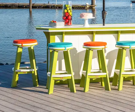 An outdoor bar on a wooden deck by the water with a white and green counter, four bar stools with orange and teal cushions, a bowl of apples, a stack of plates with strawberries, and a white cake. The background features a calm lake and a dock.
