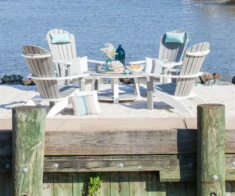 conversation-table-thumbnail.webp A beachside seating area on a wooden deck with four white Adirondack chairs around a round table, decorated with a blue vase, white flowers, and a bowl of snacks. The background features a calm lake.