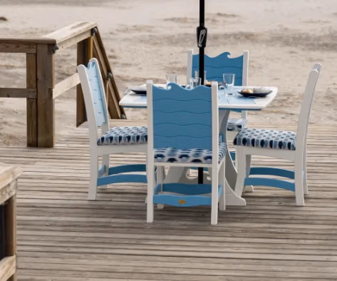 A beachside dining setup on a wooden boardwalk with a round table, four white chairs with blue cushions, and a patterned umbrella. The sandy beach and ocean waves are in the background.
