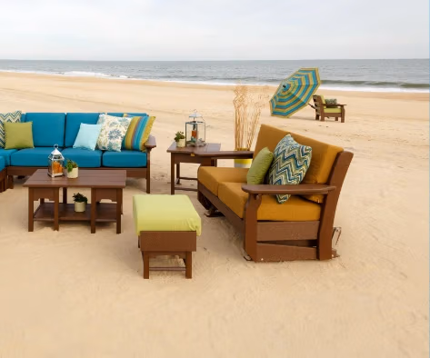 A beachfront seating area with blue and yellow cushioned sofas, a green ottoman, and wooden coffee tables. Decor includes patterned pillows, potted plants, and a lantern. A striped umbrella and chairs are in the background near the ocean.