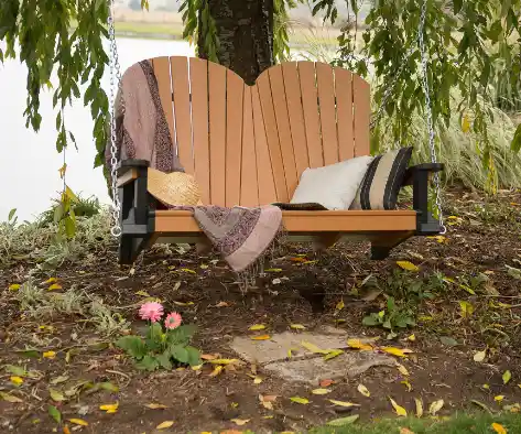 A wooden porch swing with a heart-shaped backrest hangs under a tree near a lake, decorated with a blanket, pillows, and a straw hat. Pink flowers and fallen leaves are on the ground.