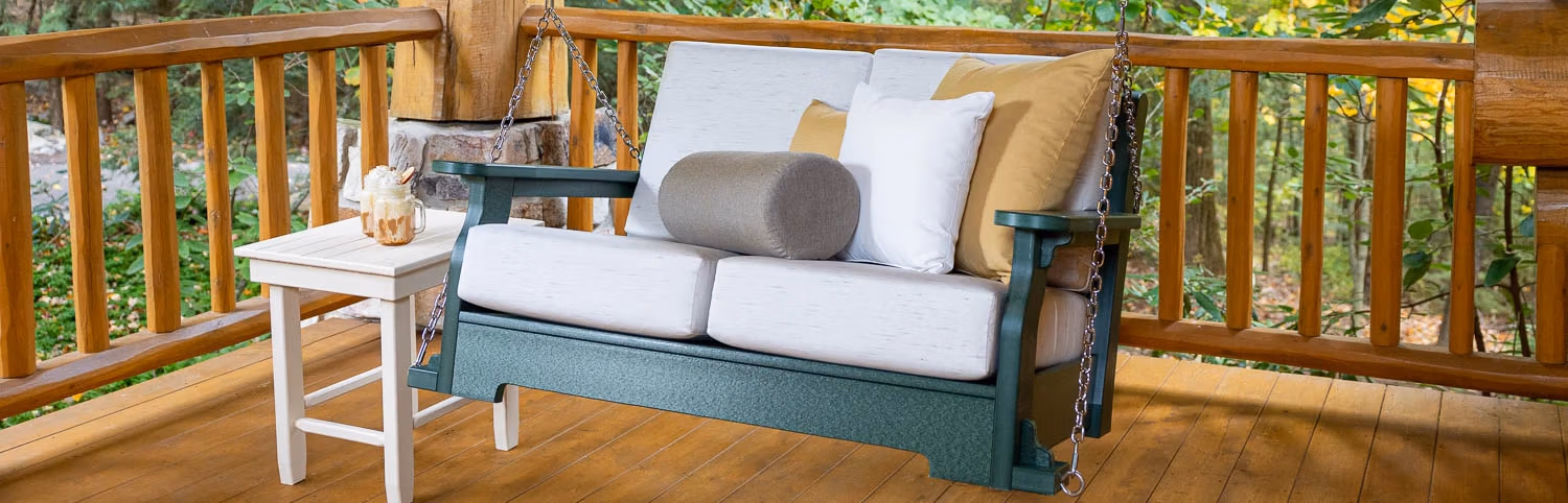 A teal poly furniture porch swing with beige cushions, decorated with gray, white, and yellow pillows, hangs on a wooden deck by a log cabin. A white poly furniture table nearby holds two drinks with whipped cream, with trees in the background.